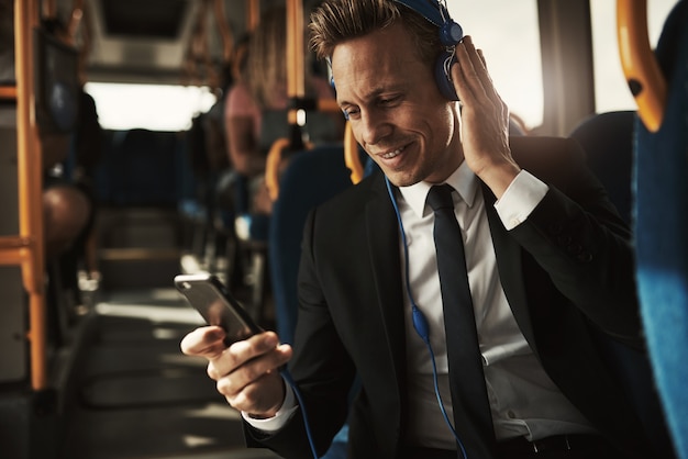 Jovem empresário sorridente de terno sentado em um ônibus ouvindo música em fones de ouvido