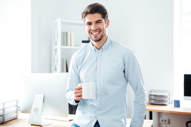 Jovem empresário sorridente em pé e bebendo café no escritório