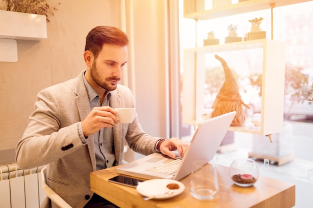 Jovem empresário sorridente em uma pausa em um café. Ele está bebendo café e trabalhando no laptop.