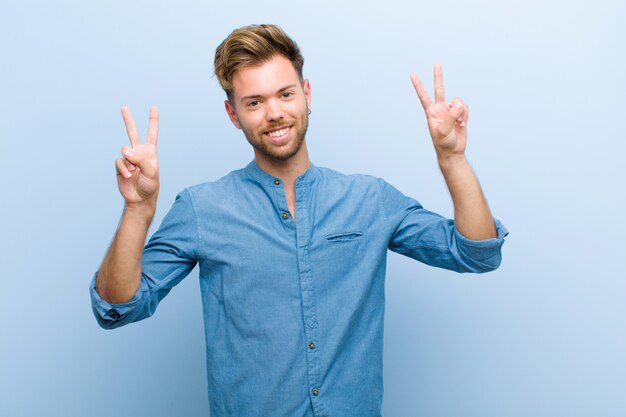 Jovem empresário sorrindo e olhando feliz, amigável e satisfeito, gesticulando vitória ou paz com as duas mãos contra o fundo azul