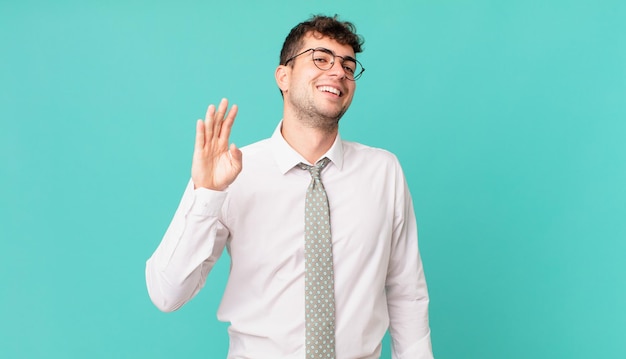 Jovem empresário sorrindo feliz e alegre, acenando com a mão, dando as boas-vindas e cumprimentando você ou dizendo adeus