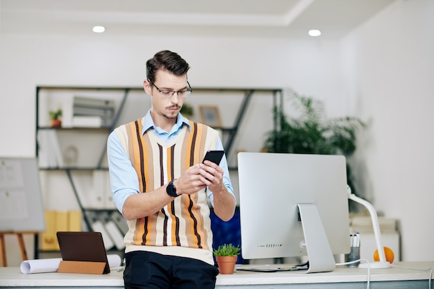 Jovem empresário surpreso com colete listrado em pé em sua mesa de escritório e verificando mensagens de texto no smartphone