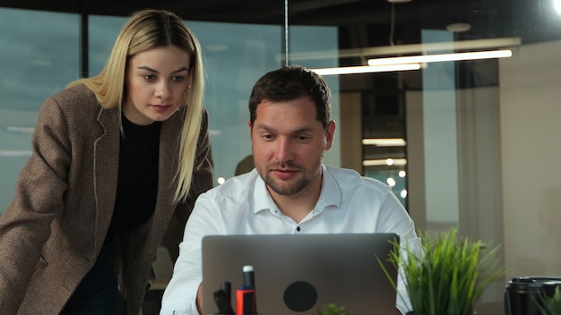 Jovem empresário trabalhando em um laptop na mesa em um escritório de plano aberto moderno, sendo acompanhado por uma colega para uma reunião informal