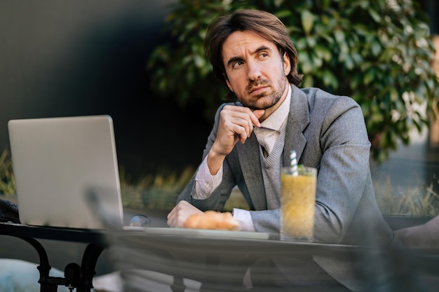 Jovem empresário trabalhando no laptop e pensando em algo enquanto está sentado no café ao ar livre
