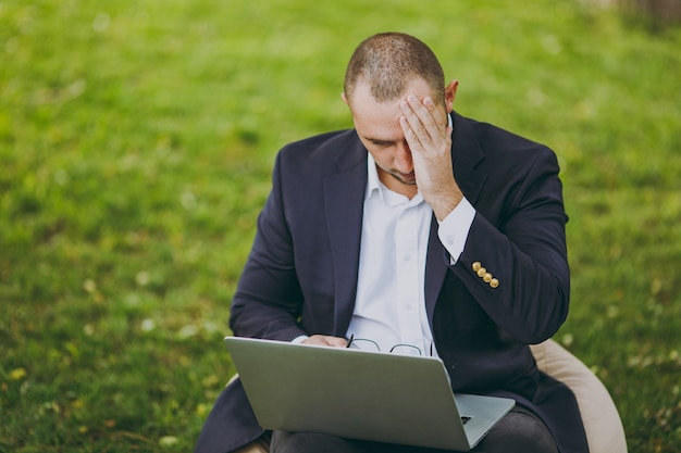 Jovem empresário triste e cansado em camisa branca, terno clássico, óculos. Homem sente-se no pufe macio, se preocupa com os problemas, trabalha no computador laptop pc no parque da cidade, no gramado verde ao ar livre. Conceito de escritório móvel.