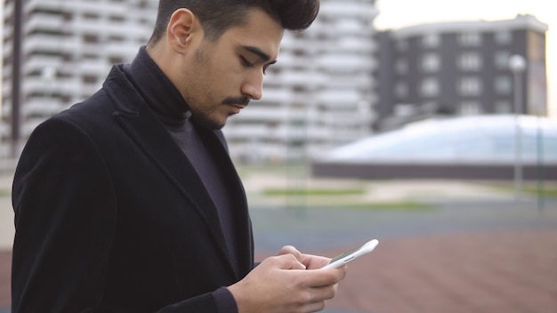 Jovem empresário urbano profissional em terno preto, andando na rua usando a mensagem de sms de mensagens de texto do app no smartphone.