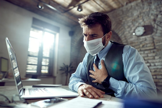 Jovem empresário usando máscara facial e sentindo dor no peito enquanto trabalhava no laptop no escritório durante a epidemia de vírus
