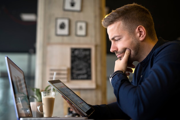 Jovem empresário usando tablet e laptop em um café, analisando projetos.