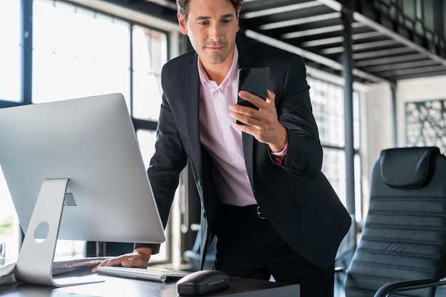 Jovem empresário vestindo terno preto em pé perto da mesa com computador sorrindo olhando para o telefone Gerente de escritório com telefone trabalhando no escritório moderno com janelas de vidro fundo desfocado