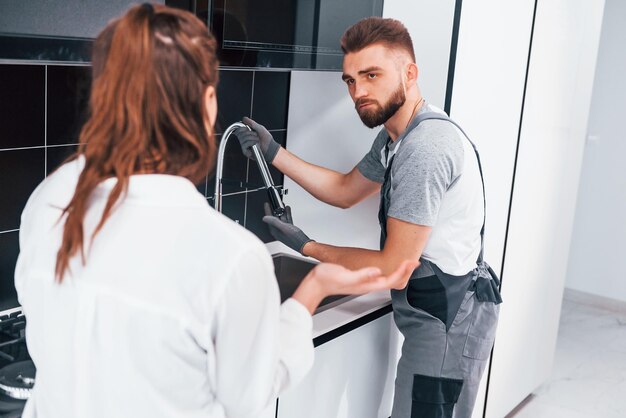 Jovem encanador profissional de uniforme cinza mostra resultados de trabalho para dona de casa na cozinha