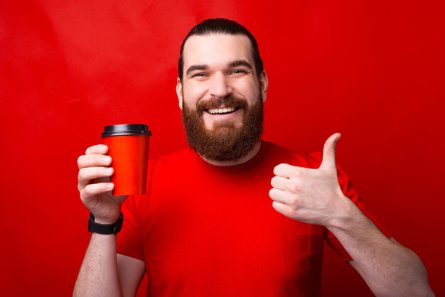 Jovem encantador com barba aparecendo o polegar e segurando o copo de papel vermelho com café para viagem