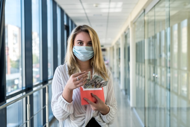 Jovem encantadora com uma máscara médica segurando um envelope com dinheiro no shopping. conceito de finanças