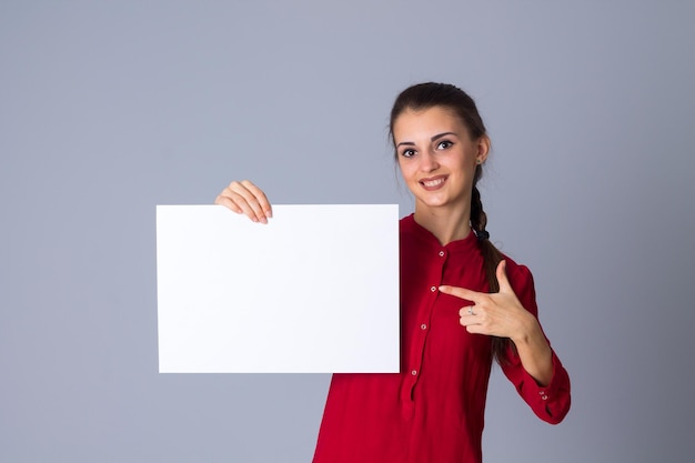 Jovem encantadora de blusa vermelha segurando uma folha de papel branca sobre fundo cinza no estúdio