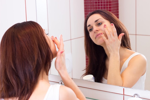 Jovem encantadora de camisa usando creme facial na frente do espelho em seu banheiro