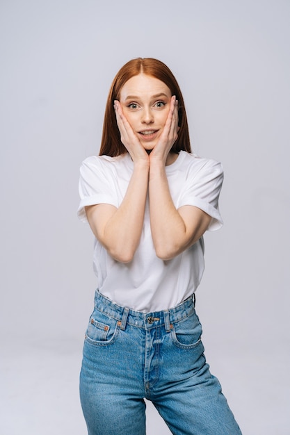 Jovem encantadora espantada usando camiseta e calça jeans sorrindo com as mãos na bochecha