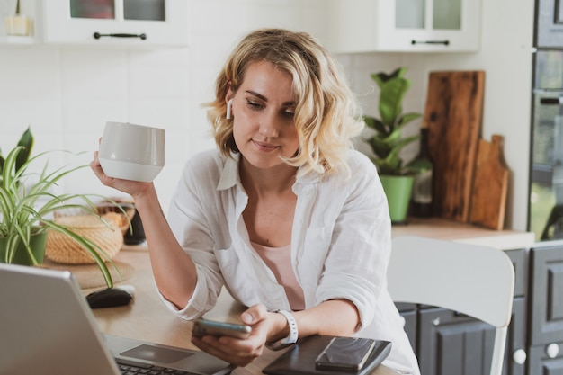 Jovem encantadora na cozinha bebe café e usa smartphone