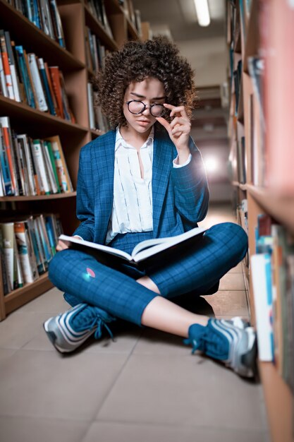 Jovem encaracolada linda de óculos e terno azul, sentado com os livros na biblioteca.