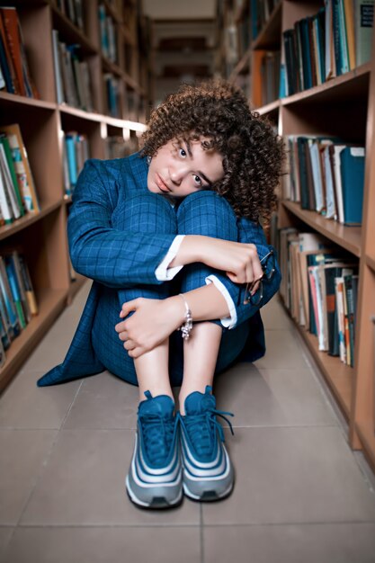 Jovem encaracolada linda de óculos e terno azul, sentado com os livros na biblioteca.