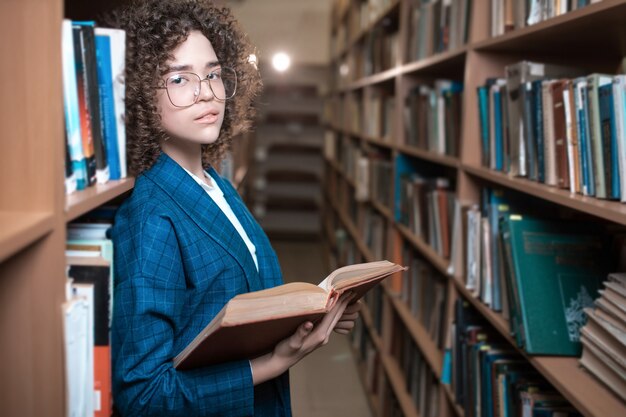 Jovem encaracolada linda de óculos e um terno azul está de pé na biblioteca