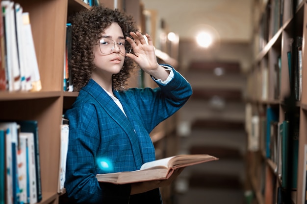 Foto jovem encaracolada linda de óculos e um terno azul está de pé na biblioteca