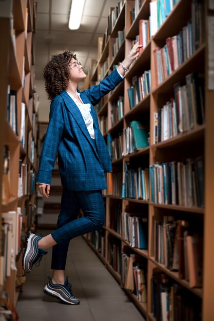 Jovem encaracolada linda de óculos e um terno azul está de pé na biblioteca