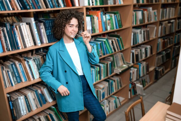 Jovem encaracolada linda de óculos e um terno azul está de pé na biblioteca