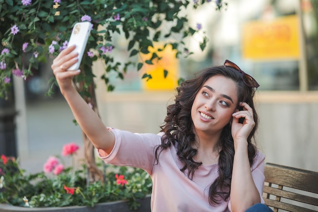 Jovem encaracolada usa um telefone para redes sociais Um modelo tira uma selfie na rua de uma cidade europeia no verão Chamada de vídeo