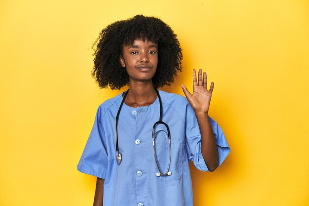 Jovem enfermeira afro-americana em estúdio com fundo amarelo sorrindo alegremente mostrando o número cinco