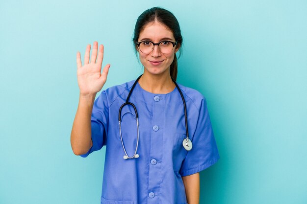 Jovem enfermeira caucasiana isolada em um fundo azul, sorrindo alegre mostrando o número cinco com os dedos.