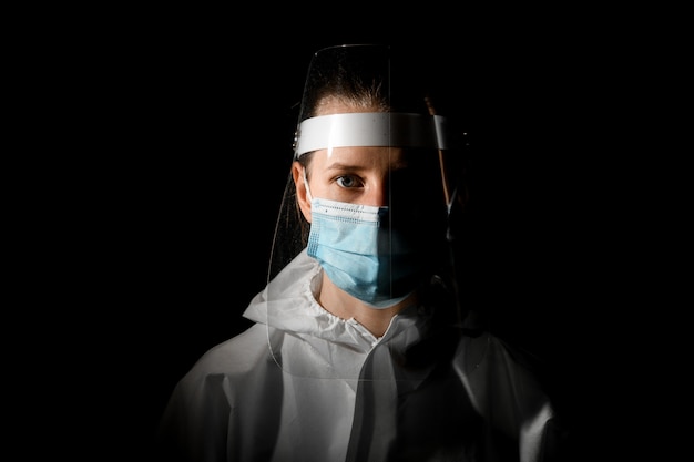 Jovem enfermeira feminina na máscara médica e escudo protetor na cabeça no quarto escuro