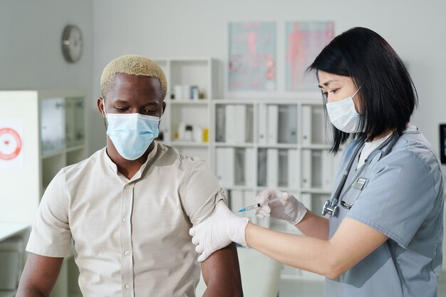 Jovem enfermeira morena de etnia asiática em uniforme e máscara protetora vacinando um cara africano sentado em consultório médico de clínicas modernas