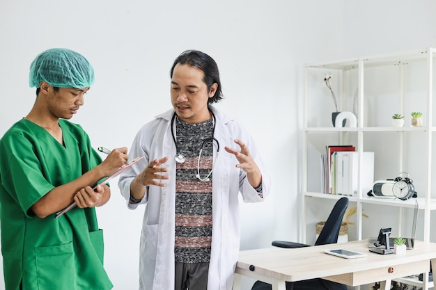 Jovem enfermeira vestindo uniforme de cirurgia, auxiliando o médico a preencher o registro médico na clínica.