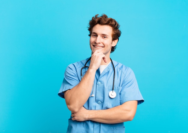 Jovem enfermeiro sorrindo com uma expressão feliz e confiante com a mão no queixo, pensando e olhando para o lado