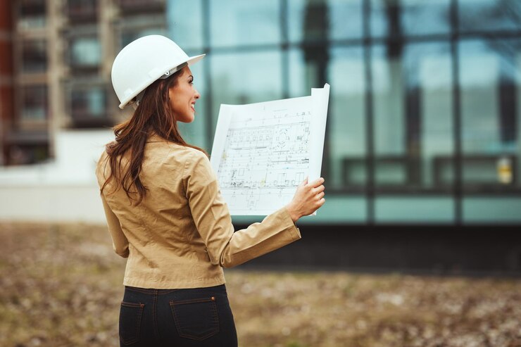 Foto jovem engenheira bem sucedida com capacete branco e plantas nas mãos no canteiro de obras analisando novo projeto.