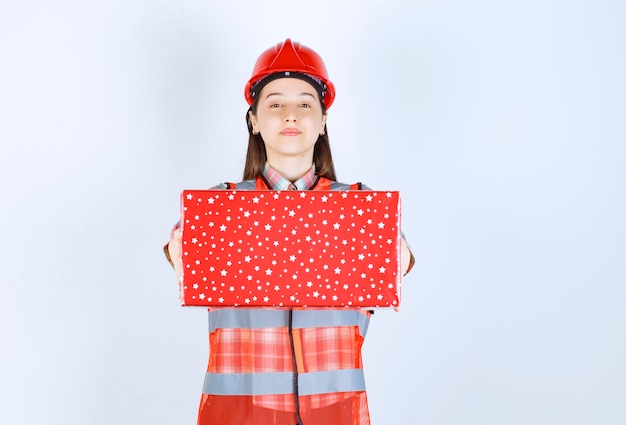 Foto jovem engenheira no capacete vermelho, mostrando a caixa de presente vermelha.