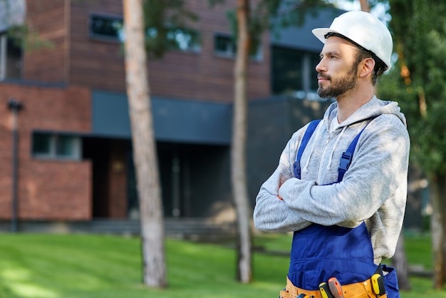 Jovem engenheiro barbudo com capacete olhando para longe, posando ao ar livre enquanto trabalhava na construção de uma casa de campo