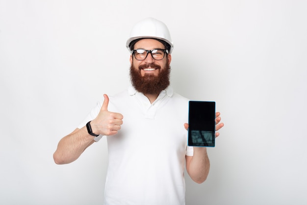 Jovem engenheiro barbudo está segurando um tablet e aparecendo o polegar.