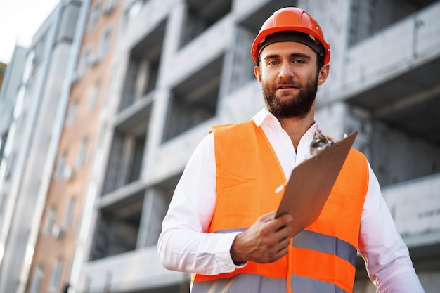 Jovem engenheiro em workwear parado no canteiro de obras com a prancheta