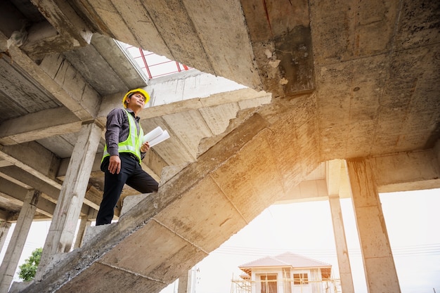 Jovem engenheiro profissional trabalhador em capacete protetor e papel de plantas disponíveis no local de construção do edifício da casa