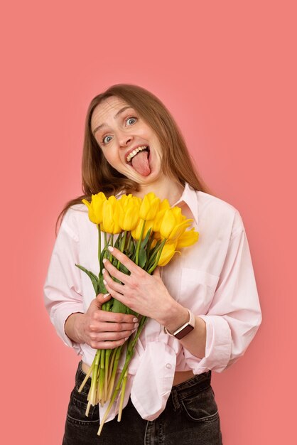 Jovem engraçada segurando buquê de flores amarelas e mostrando a língua para a câmera Garota com tulipas faz caretas Isolado em fundo rosa Moldura vertical