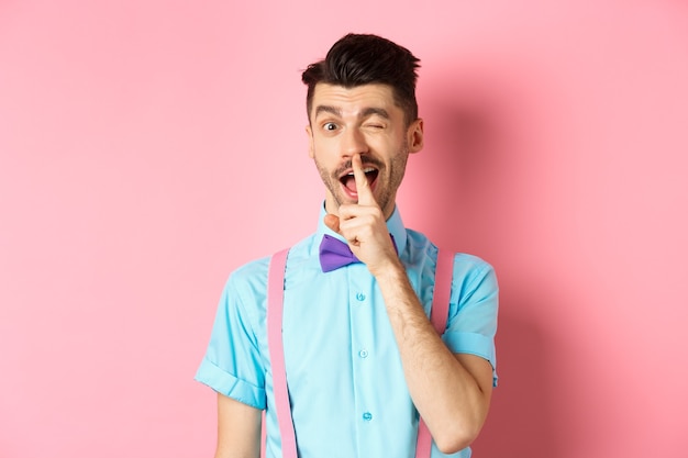 Jovem engraçado planejando uma surpresa, silenciando e piscando para a câmera, pedindo para ficar quieto, contando um segredo, pisando em rosa.