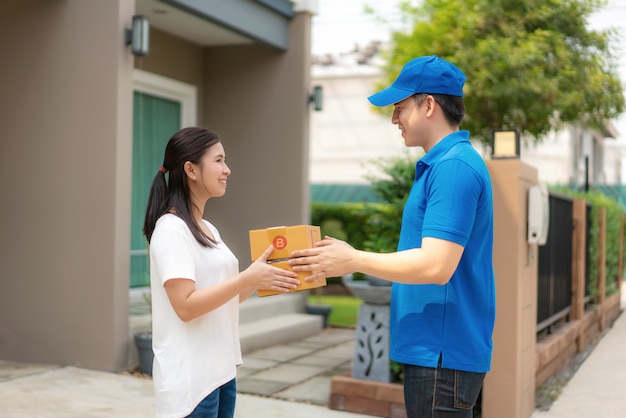 Jovem entrega asiática no sorriso uniforme azul e segurando a pilha de caixas de papelão na frente de casa