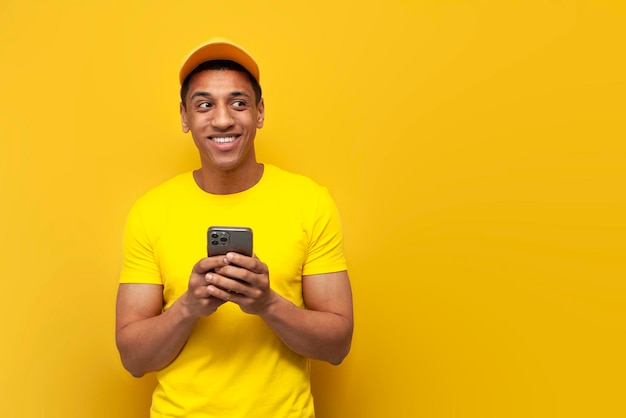 Jovem entregador afro-americano de uniforme amarelo usando smartphone e olhando para o espaço da cópia