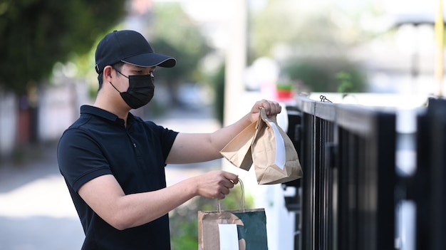 Jovem entregador asiático na máscara protetora, segurando o saco de papel com comida e esperando o cliente na porta de casa.