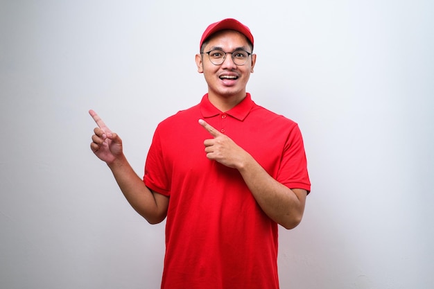 Jovem entregador chinês bonito vestindo boné de pé sobre fundo branco isolado com um grande sorriso no rosto, apontando com a mão e o dedo para o lado, olhando para a câmera