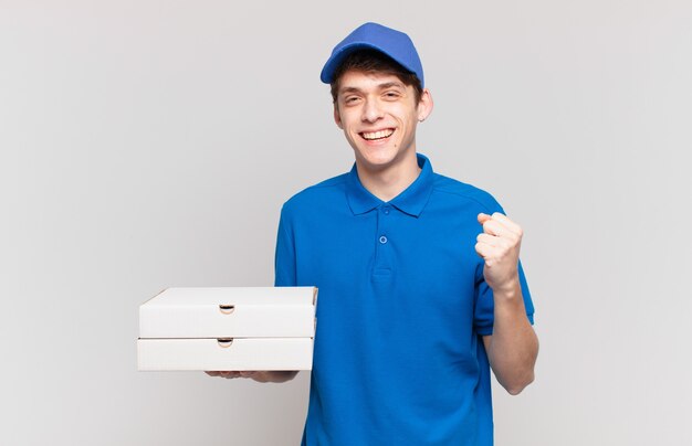 Jovem entregador de pizza se sentindo chocado, animado e feliz, rindo e comemorando o sucesso, dizendo uau!
