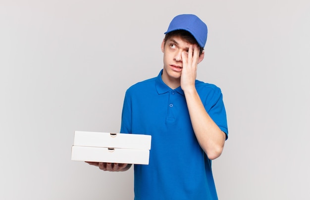 Jovem entregador de pizza se sentindo entediado, frustrado e com sono depois de uma tarefa cansativa, enfadonha e tediosa, segurando o rosto com a mão