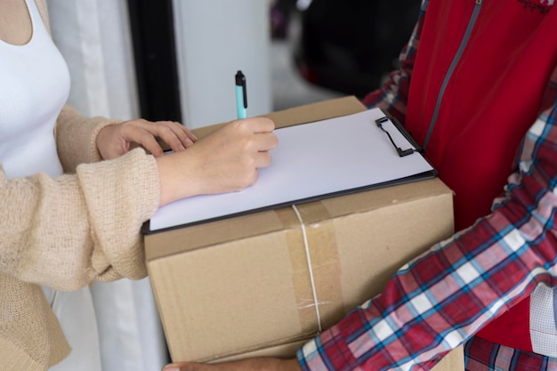 Jovem entregador de uniforme vermelho segurando uma caixa de papelão entregando pacote e mulher colocando assinatura no documento