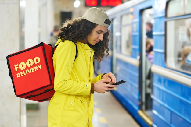 Jovem entregador em mensagens de texto de uniforme amarelo ou rolagem em smartphone na estação de metrô