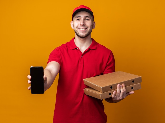 Jovem entregador sorridente, usando uniforme com tampa, segurando caixas de pizza e mostrando o telefone